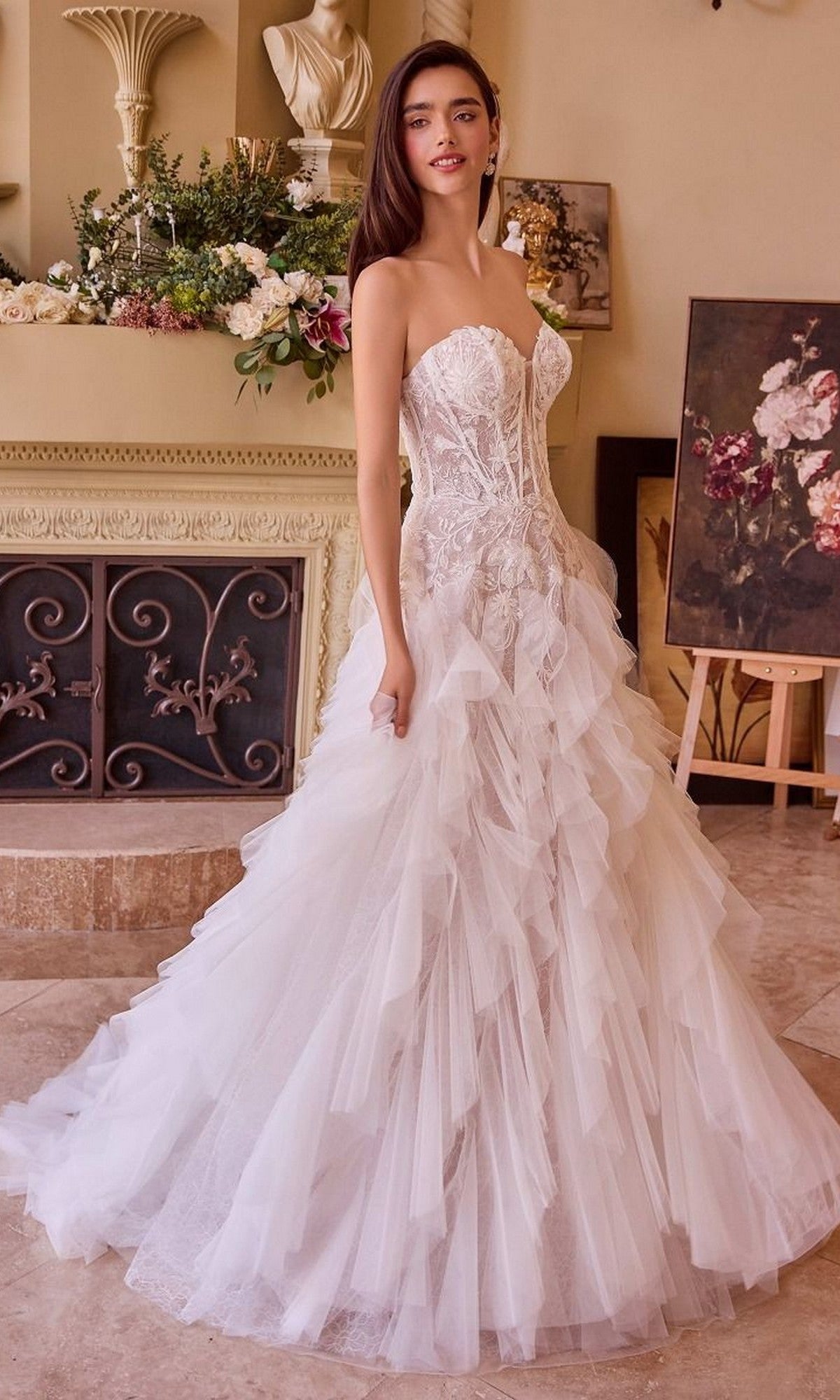 Indoors, a woman wears the Andrea and Leo WL016 long wedding dress with a strapless, lace-applique corset bodice and a ruffle tulle A-line skirt. Floral arrangements and artwork decorate the background.